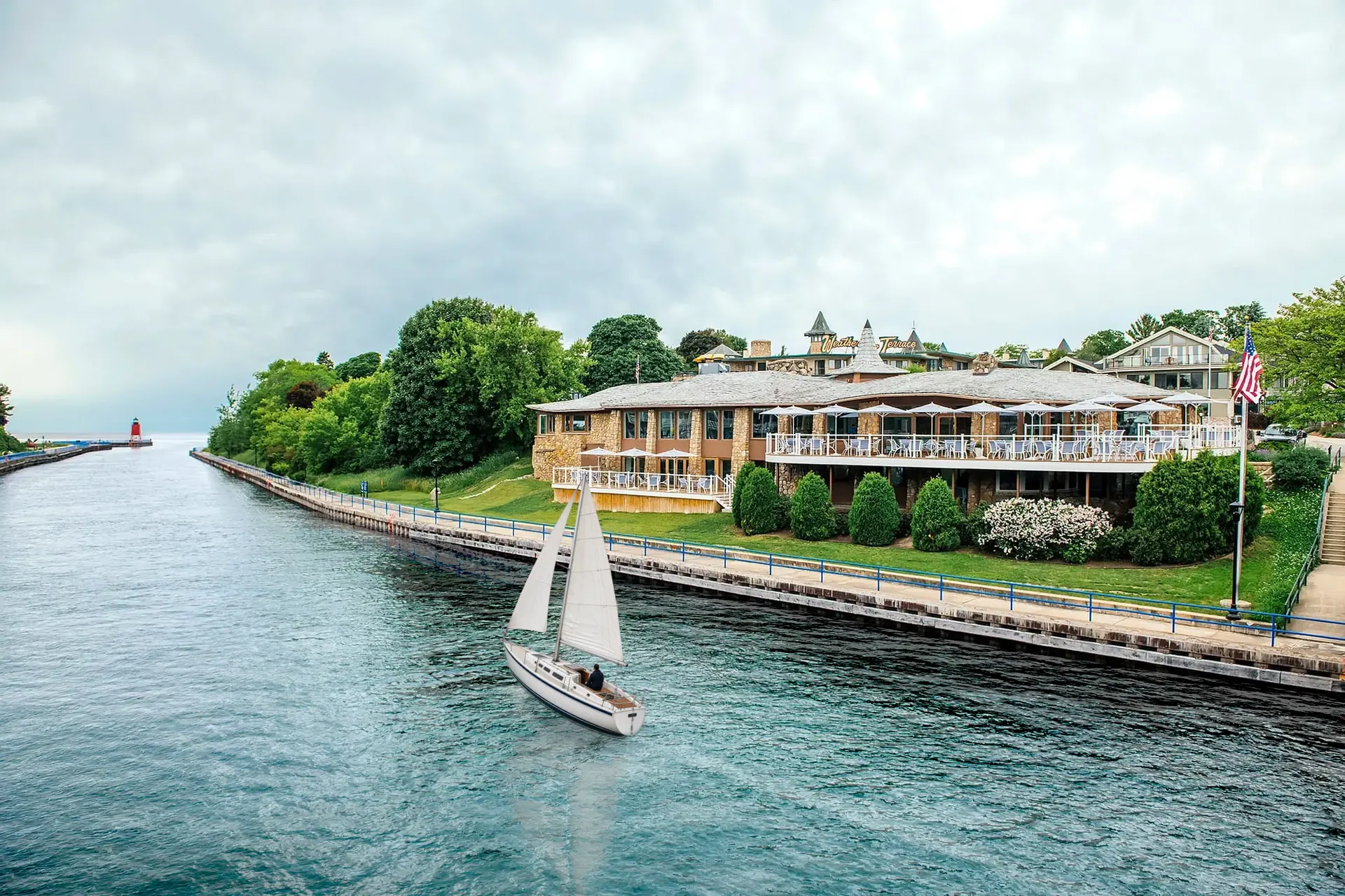 The Weathervane's stone building on its lawn beside the Pine River Channel, with the blue seawall railing and the red pierhead lighthouse in the distance, and a sailboat heading out the channel toward Lake Michigan